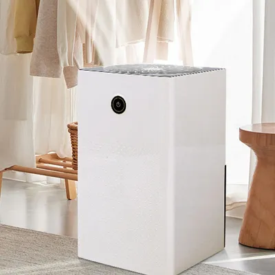 A white dehumidifier is placed on a rug in a room with clothes hanging in the background and a wooden stool to the side.