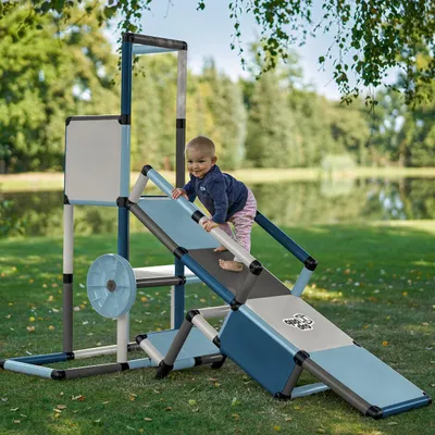A baby is playing on a colorful playground structure in a grassy outdoor area with a lake in the background.