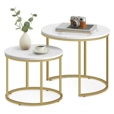 A set of two round nesting coffee tables with white marble-effect tops and gold metal legs, styled with a vase of greenery and a coffee cup on the smaller table, and a book and camera on the larger table.