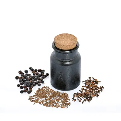 A small dark ceramic spice jar with a cork stopper is shown in the center, surrounded by piles of juniper berries, cumin seeds, and cloves.