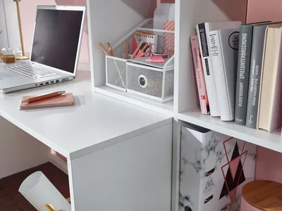 A close-up view of a white desk with a laptop, stationery, and books on shelves.