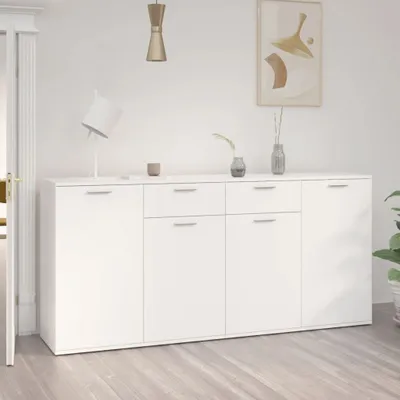 A white sideboard with drawers and doors is shown in a room with a light wooden floor and white walls. A lamp, a small cup, and two vases with plants are on top of the sideboard. A framed picture hangs on the wall above the sideboard.
