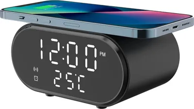 A black digital alarm clock with a wireless charging pad on top, charging a smartphone. The clock displays the time and temperature.