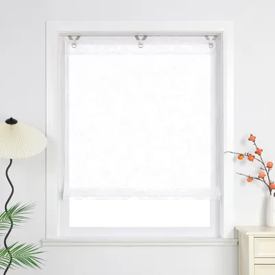 A white patterned roller blind is shown in a window frame, with a lamp and plant on the left and a vase with branches on the right.