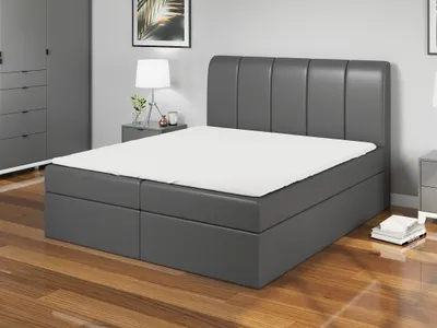 A dark gray boxspring bed with a white mattress and a headboard with vertical panels, set in a bedroom with a wardrobe and a nightstand.
