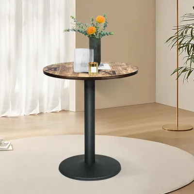 A round bistro table with a wood-look top and a black pedestal base, set on a cream rug in a room with sheer curtains and a plant.