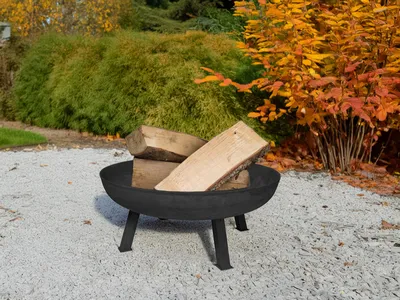 A black metal fire pit filled with logs sits on a gravel surface in a garden setting with autumn foliage in the background.