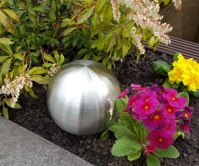 A decorative metal sphere sits in a garden bed surrounded by flowers and foliage.