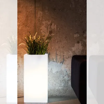 A lit white planter with green grass inside, placed next to a dark sofa against a textured concrete wall.