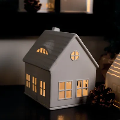 A ceramic house-shaped lantern with illuminated windows sits on a dark surface, with a pinecone in the foreground and another lantern in the background.