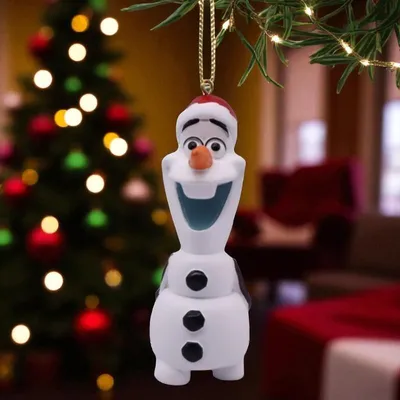 An Olaf Christmas ornament hangs from a tree branch, with a decorated Christmas tree and a blurred room in the background.