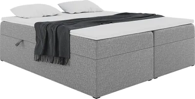 A gray upholstered box spring bed with two white pillows and a dark gray blanket draped over it, shown from a slightly elevated side angle against a white background.