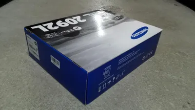 A blue and white cardboard box for a Samsung toner cartridge sits on a gray surface.