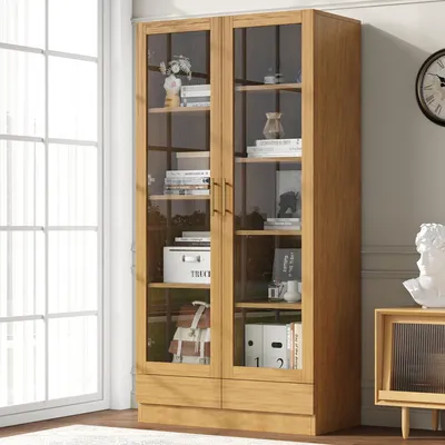 A wooden display cabinet with glass doors filled with books and decorative items, placed near a window and a wall clock.