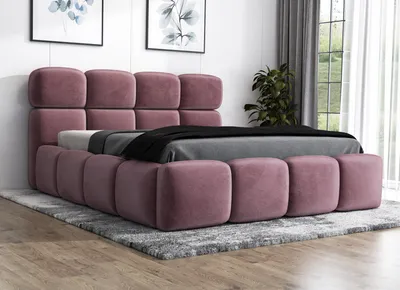 A pink upholstered bed with a tufted headboard and frame, placed on a gray rug in a room with artwork on the wall and a plant in the background.