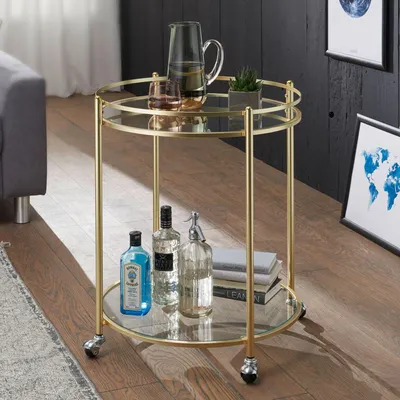 A gold bar cart with glass shelves is shown in a room with a sofa and framed pictures on the wall.