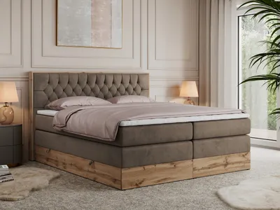A bedroom scene featuring a large boxspring bed with a tufted headboard and a wooden base, set against a neutral-toned wall with decorative molding. A bedside table with a lamp is visible to the left, and a large window with curtains and a plant is on the 