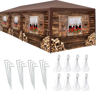 A long, brown gazebo with a printed image of a wooden chalet facade, including windows and stacked firewood. Below the gazebo are metal stakes and white ropes, likely accessories for assembly.