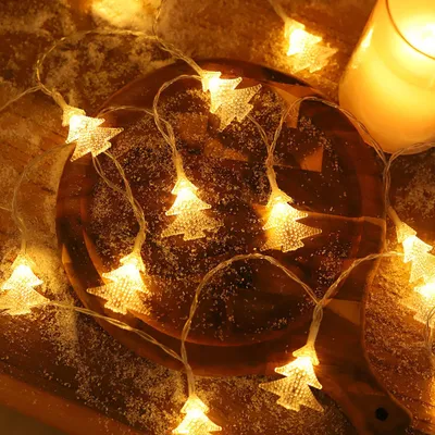 A string of Christmas tree-shaped lights is draped over a wooden surface dusted with artificial snow, with a lit candle in the background.
