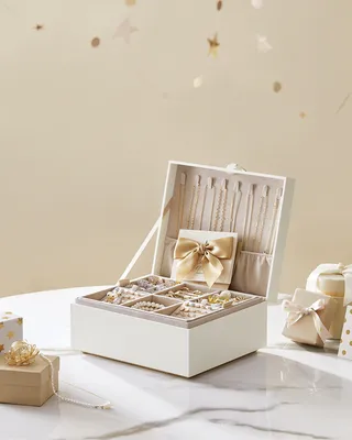A white jewelry box with multiple compartments is open, displaying various jewelry pieces. The box is placed on a marble surface, with gift boxes and decorative elements in the background.