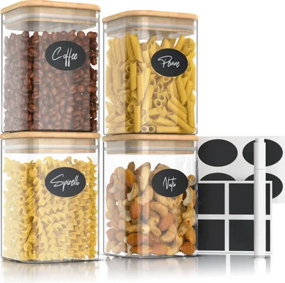 Four clear glass storage jars with bamboo lids, filled with coffee beans, pasta, fusilli, and nuts. Each jar has a black oval label with white text indicating its contents.