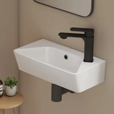 A modern white ceramic wall-mounted sink with a black faucet is shown against a light beige wall. A small potted plant and a bottle are visible on a wooden stool to the left.