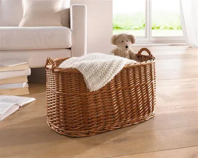 A wicker basket with a blanket and a teddy bear sits on a wooden floor next to a sofa. A window with greenery is visible in the background.