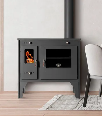 A dark gray wood-burning stove with a glass door showing flames, a glass oven door, and a metal chimney pipe, set against a light gray and white textured wall with a wooden floor and a rug.