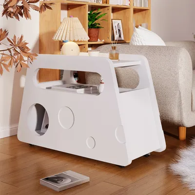 A white side table with circular cutouts is placed next to a sofa in a living room setting. A bookshelf is visible in the background.
