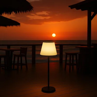 A solar-powered floor lamp is illuminated on a wooden deck at sunset, with the ocean and a bar in the background.
