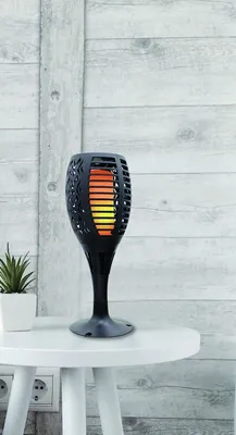 A black solar-powered garden torch light with flickering LED flames sits on a white side table, next to a small potted plant. The background is a light gray wooden wall.