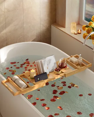 A relaxing bath setup with rose petals, a book, wine, and bath accessories on a bathtub tray.