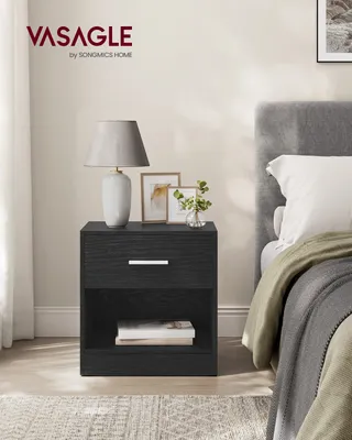 A black nightstand with a drawer and an open shelf is placed next to a bed.