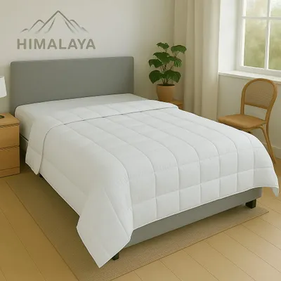 A white comforter is laid out on a bed with a grey headboard, next to a nightstand and a plant.