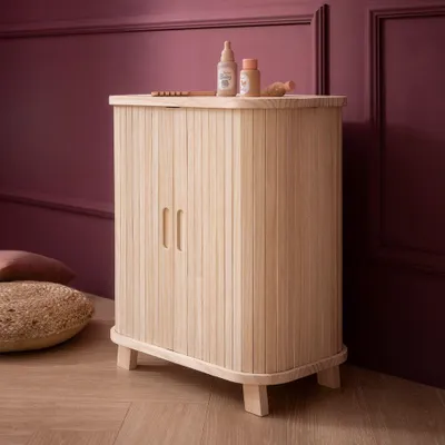 A wooden cabinet with two doors and small legs, placed on a wooden floor next to a wall painted in a deep purple color. There are two small bottles and a toy on top of the cabinet.