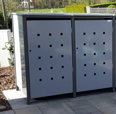 Two metal garbage bins are shown outdoors on a paved area, with a garden and fence in the background.