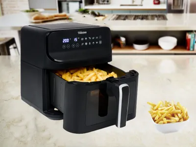 A black air fryer filled with french fries is shown on a kitchen counter, with a bowl of french fries next to it. The background shows a kitchen setting.