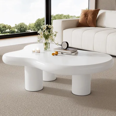 A white, irregularly shaped coffee table is placed in a modern living room setting, with a sofa and a large window in the background.