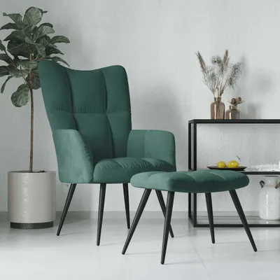 A dark green velvet armchair and ottoman are displayed in a minimalist room setting with a potted plant and a shelf with decorative items.