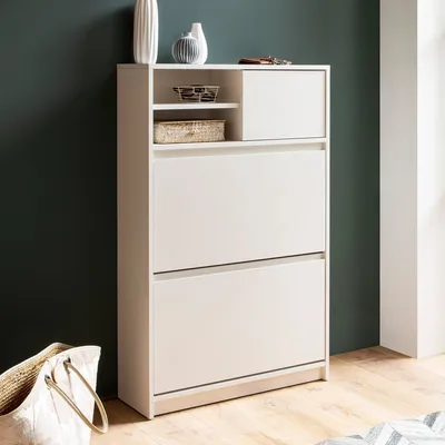 A white shoe cabinet with two flip-down drawers and a small shelf with two baskets on top, placed against a dark green wall.