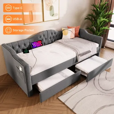 A gray upholstered daybed with two pull-out drawers, set up in a room with decorative pillows and a plant. The bed has a tablet and a charging device on it, and storage pockets on the side.