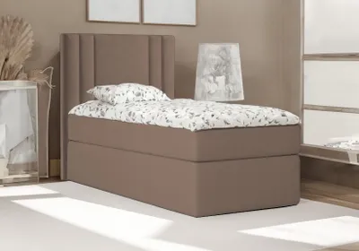 A single bed with a brown upholstered base and headboard, dressed with patterned bedding and a pillow. The bed is placed in a room with a neutral-toned wall and wooden floor. A decorative lamp and a framed picture are visible in the background.
