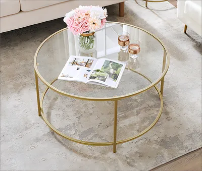 A round glass coffee table with a gold metal frame, decorated with a vase of flowers and two glasses of drinks.
