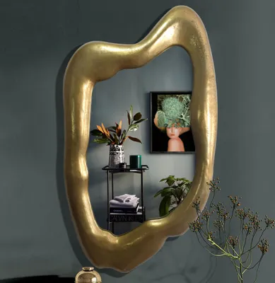 A large, irregularly shaped gold mirror hangs on a dark gray wall, reflecting a small shelf with decorative items and a framed picture.