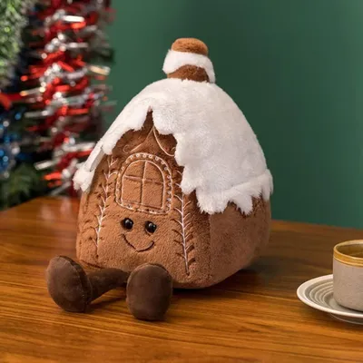 A plush gingerbread house toy sits on a wooden table, with Christmas tinsel in the background.