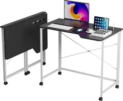 A black folding desk with white metal legs and wheels, shown next to a standard computer desk with a laptop, tablet, keyboard, mouse, and coffee cup on it.
