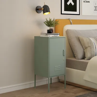 A mint green nightstand with a locked door and slatted ventilation is placed next to a bed with yellow and white bedding. A wall lamp with a black shade and gold accents is mounted on the wall above the nightstand. Books and a small potted plant are on top