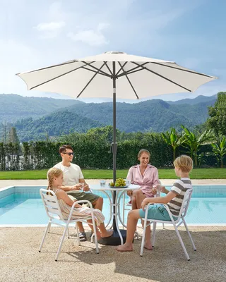 A family is enjoying a meal under a large umbrella by a swimming pool with a mountainous backdrop.