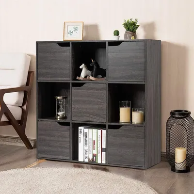 A gray wooden bookshelf with 9 compartments, some of which have doors, is shown in a room with a chair and a rug.