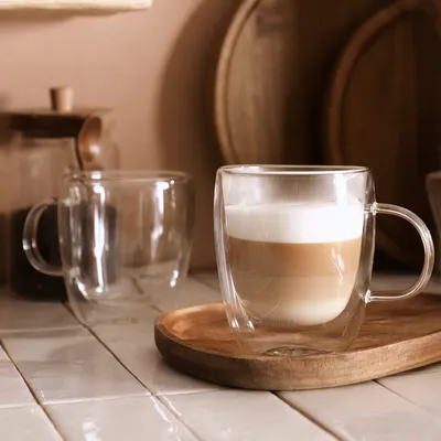 Two double-walled glass mugs, one filled with a layered coffee drink, are placed on a wooden tray on a tiled surface. A jar and wooden plates are in the background.